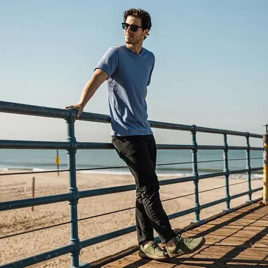 Man leaning on walkway railing at the beach wearing casual clothes and olive green barefoot sneakers.