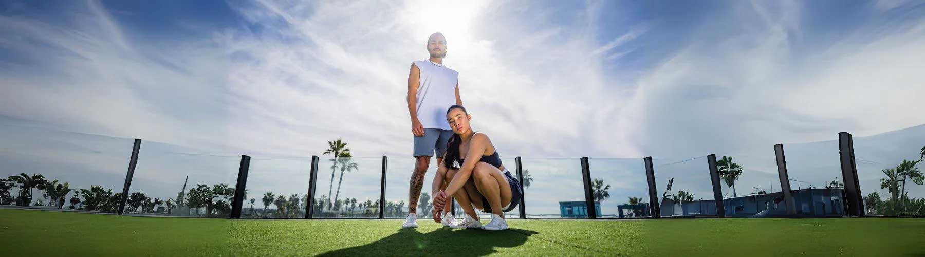 Wide view image of two people outside on green grass and the sky behind them, wearing sport clothes and lightweight barefoot athletic shoes.