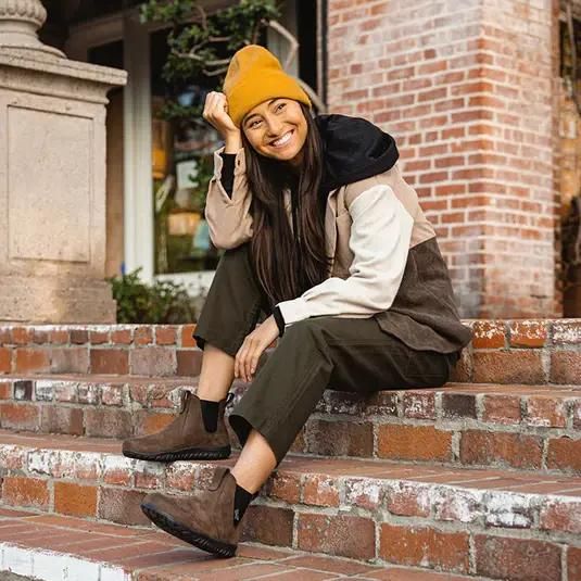 Women sitting on brick stairs outside in winter clothes, wearing waterproof brown leather barefoot Lems Chelsea boots.