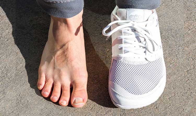 A white Lems barefoot Primal Zen shoe next to a bare foot on a concrete surface, to demonstrate the foot-shaped barefoot shoe with a wide toe box.