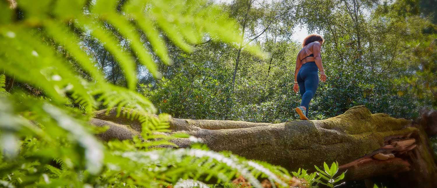 Person walking through a forest with large trees and ferns, wearing active wear and flexible, lightweight barefoot shoes.