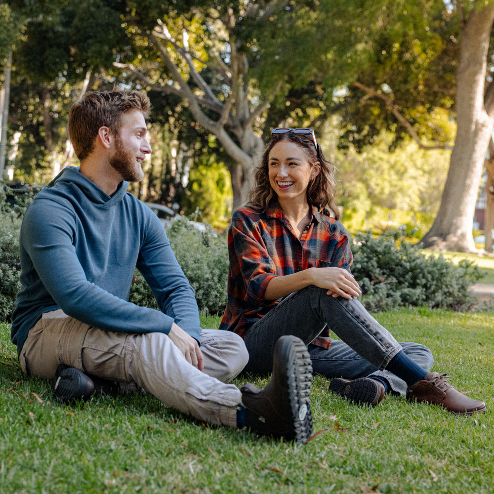 Two people sitting on grass in a park, one wearing rugged barefoot leather boots and the other wearing barefoot leather lace-up shoes.