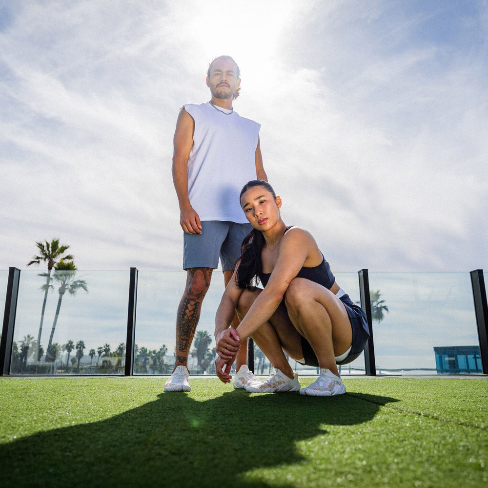 Two people posing outdoors with palm trees and a clear sky in the background, wearing sport clothes and lightweight barefoot athletic shoes.