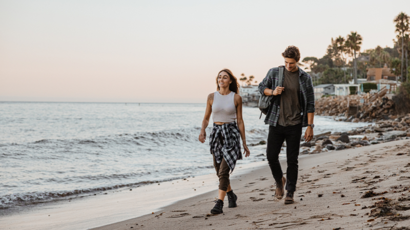 Two people walking along the beach at sunset wearing Lems barefoot shoes, featuring casual outdoor styling, natural movement, and minimalist comfort by the coastline.