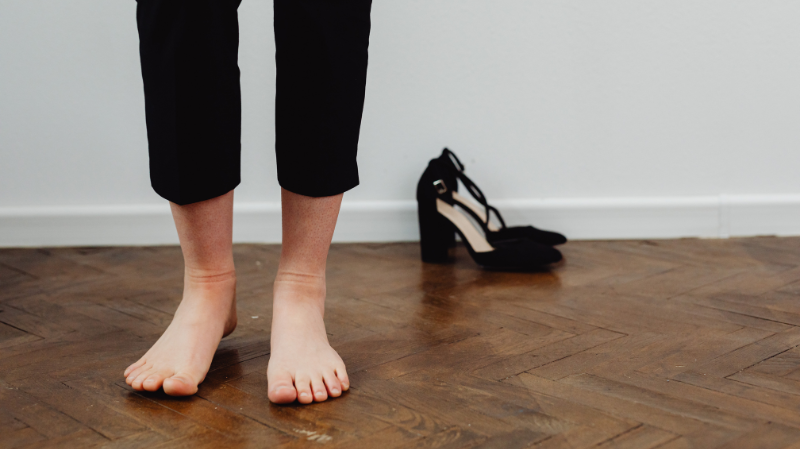 bare feet standing on wooden floor with narrow high heels in background highlighting contrast with restrictive footwear