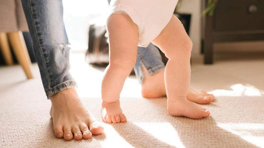 Baby taking first steps barefoot on a carpet next to an adult, highlighting natural foot movement and development