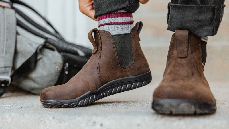 Lems brown leather Chelsea waterproof barefoot boots with flexible barefoot sole being worn on concrete steps, featuring elastic side panels and a rugged outsole.