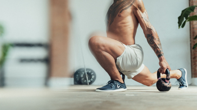 Athletic man performing a kettlebell exercise indoors while wearing Vivobarefoot barefoot shoes, demonstrating strength and balance during a workout.