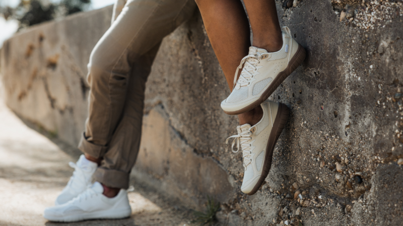 Two people sitting against a rock wall outside, wearing zero drop minimalist shoes with flexible soles for natural movement.