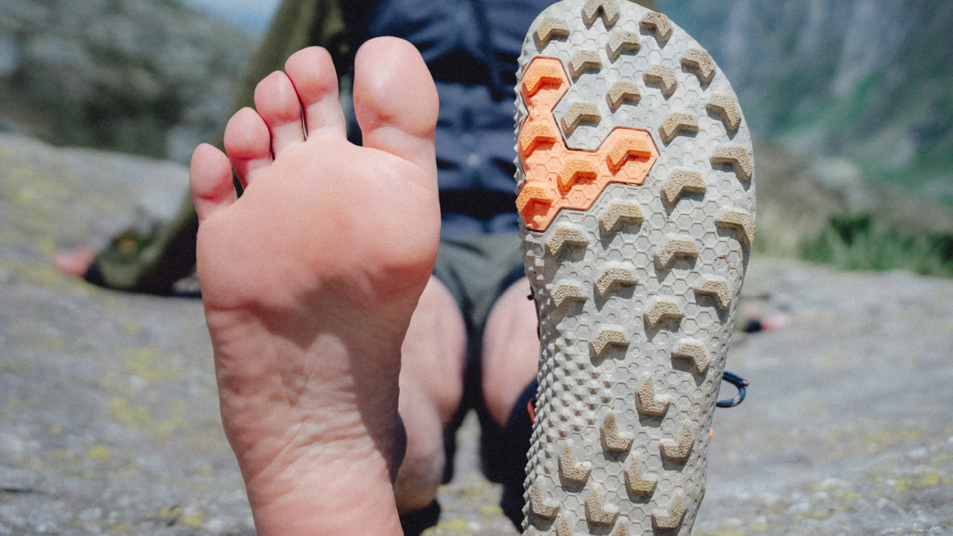 A man sitting on a rocky surface during a hike wearing no shoe on the right foot, and a zero drop, wide toe box minimalist shoe on the left foot to show the foot-shaped design of barefoot shoes.