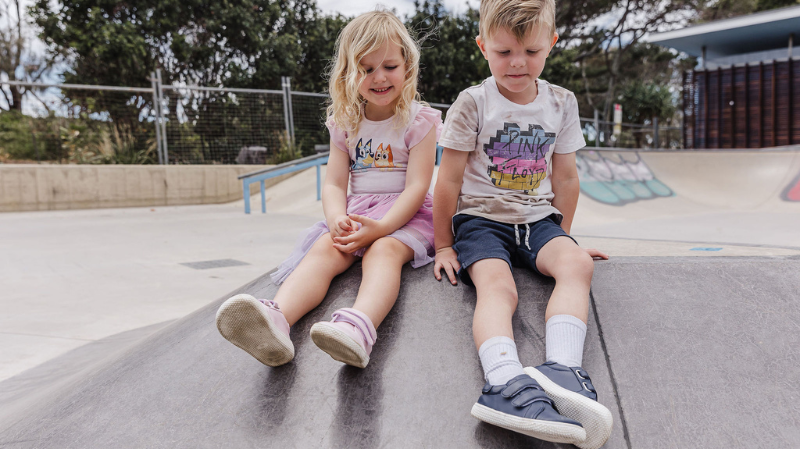 A young girl and boy sitting on a playground wearing barefoot kids shoes with velcro straps