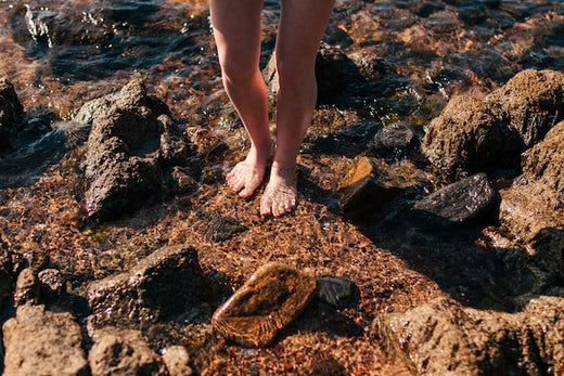Person standing barefoot in shallow water among rocks at the shoreline, with small waves moving around their feet.