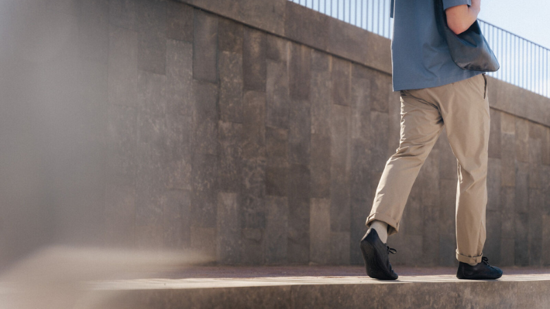 A man walking to work outside wearing black men’s barefoot business shoes with a wide toe box and zero drop sole, dressed in smart casual clothes.