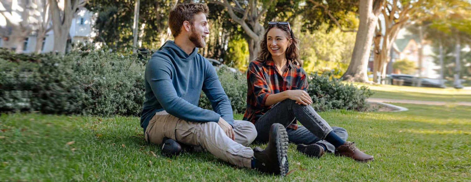Two people sitting on grass in a park outside, one wearing rugged barefoot boots and the other wearing barefoot lace-up leather shoes.